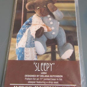 May include: A pattern for an 11-inch jointed bear in a sleeper, titled "SLEEPY." The bear is brown and blue, with a patchwork quilt. The text reads "DESIGNED BY DELANA HUTCHISON" and "INDYGO JUNCTION, INC."