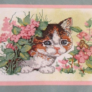 May include: Cross-stitch of a kitten with brown, white, and orange fur, surrounded by pink flowers and green leaves. The kitten has blue eyes and is centered in the image. The background is a light yellow color, and the image is framed with a pink border.