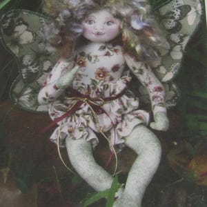 May include: A handmade fairy doll with a floral print dress and butterfly wings. The doll has a painted face, curly hair, and a ribbon belt. The wings are green with a butterfly pattern. The doll is sitting in a natural setting.
