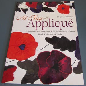 May include: A book titled "At Play with Appliqué" by Dilys A. Fronks. The cover features a white background with large red, purple, and black floral illustrations. The book details 7 template-free techniques and 10 step-by-step projects.