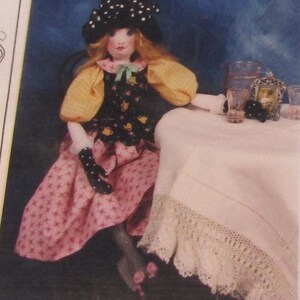 May include: A handmade doll with red hair, wearing a black hat with white polka dots, a yellow and black dress, and a pink skirt with a star pattern. The doll is seated at a table with a white tablecloth and lace trim.