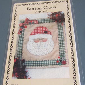 May include: A Christmas applique kit titled "Button Claus." The design features a Santa Claus face made of buttons, with a red and white checkered hat. The dimensions are 15" x 17". The kit is packaged in a clear plastic bag.