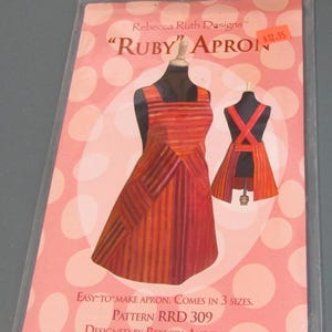 May include: A sewing pattern packet for the Ruby Apron by Rebecca Ruth Designs, featuring a front and back view of a striped orange and red apron on a pink polka dot background with the text Pattern RRD 309 and Designed by Rebecca Anderson.