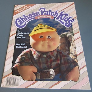 May include: A vintage Cabbage Patch Kids Magazine cover featuring a doll wearing a yellow hat with "CAT" on it. The magazine has text that reads "A Halloween Party For You" and "Hot Fall Fashions!" with a background of the Grand Canyon.