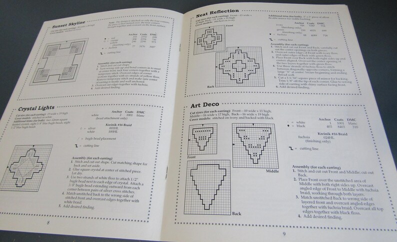 May include: An open book displaying cross-stitch patterns, such as "Sunset Skyline," "Crystal Lights," "Nest Reflection," and "Art Deco." The pages feature diagrams, instructions, and colour codes for each design.