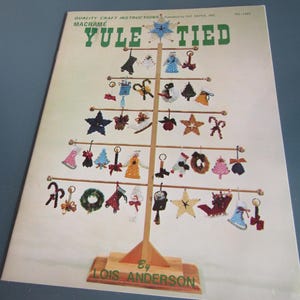May include: A vintage macrame craft instruction book titled "Yule Tied" featuring a Christmas tree design. The cover displays a wooden tree adorned with various macrame ornaments in festive colors. The author's name, Lois Anderson, is visible at the bottom.
