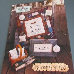May include: A craft pattern booklet titled Plain Jane Creations Old Country Stitchin featuring a patchwork quilt with snowmen, a framed embroidery piece with the text Mittens, mittens everywhere but never a pair, and a woven basket with small pillows.