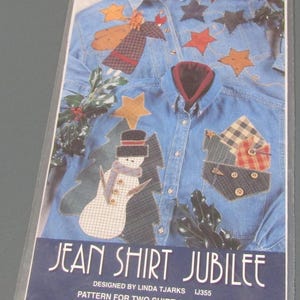 May include: A sewing pattern package titled Jean Shirt Jubilee by Indygo Junction, Inc. The cover displays two blue denim shirts decorated with fabric appliques, including a snowman, stars, and plaid patches, with the text Pattern for two shirt appliques.