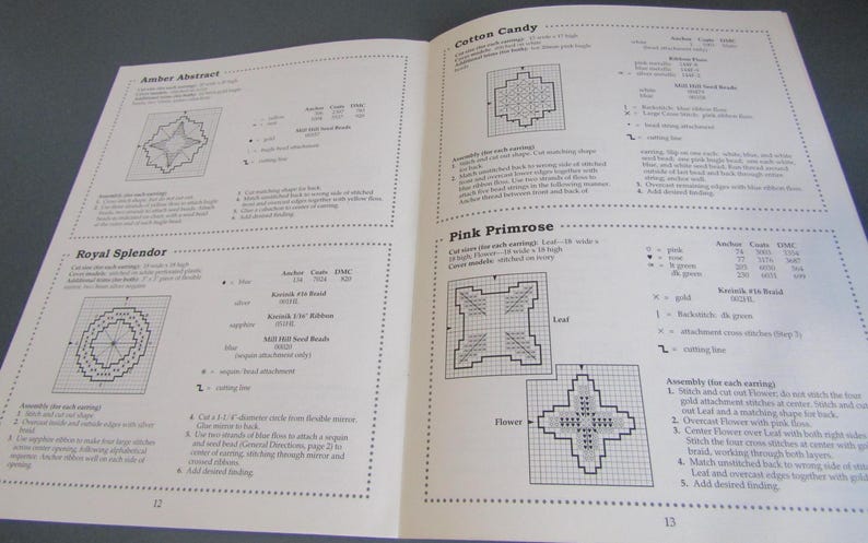 May include: Two open pages of a book featuring cross-stitch earring patterns. Designs include "Amber Abstract," "Cotton Candy," "Royal Splendor," and "Pink Primrose." Each pattern has a diagram, colour key, and instructions.