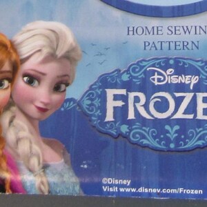 Op de afbeelding: Een blauwe en witte Disney Frozen naaipatroondoos met Anna en Elsa. De doos heeft het Disney Frozen-logo en de tekst "Home Sewing Pattern". De achtergrond toont een meerlandschap.