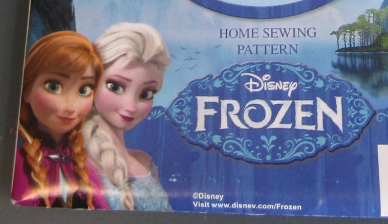 Op de afbeelding: Een blauwe en witte Disney Frozen naaipatroondoos met Anna en Elsa. De doos heeft het Disney Frozen-logo en de tekst "Home Sewing Pattern". De achtergrond toont een meerlandschap.