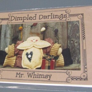 May include: A craft pattern titled "Dimpled Darlings" featuring a folk art doll named "Mr. Whimsy." The doll has a cream-colored body with a heart, and is surrounded by decorative elements. The image is framed with a decorative border.