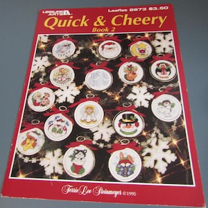 May include: A red and white book cover titled "Quick & Cheery Book 2" with various Christmas ornament designs. The ornaments are round with red bows and feature cross-stitch designs of Santa, snowmen, and angels. The cover also includes the text "Leisure Arts" and "Terrie Lee Steinmeyer ©1995."