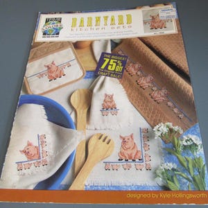 May include: A collection of kitchen textiles featuring a pig theme. Includes a potholder, placemat, napkin, and towel with cross-stitch pig designs. Wooden spoons and a blue bowl are also present. The text "BARNYARD kitchen sets" is visible.