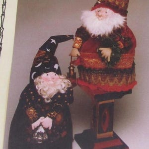 May include: Two decorative Santa Claus figures. One is standing on a pedestal, wearing a red and gold outfit with a white beard and holding a lantern. The other is a smaller figure in a black robe with gold stars and a matching hat.