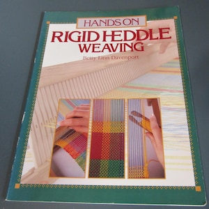 May include: A softcover instructional book titled Hands On Rigid Heddle Weaving by Betty Linn Davenport. The cover features a dark green border and three inset photographs showing a wooden loom with colorful warp and weft threads in progress.
