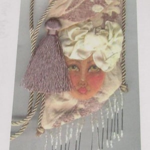 May include: A rectangular fabric pouch featuring a printed face with a cream-colored headwrap, accented by a mauve tassel, a twisted rope strap, and a fringe of clear beads hanging from the bottom edge against a patterned mauve and cream background.