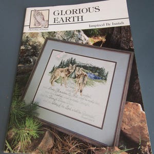 May include: A cross-stitch pattern titled "Glorious Earth" with the text "Praying Hands" and "Inspired by Isaiah." The framed artwork depicts two wolves in a forest scene. The pattern includes text from Isaiah.