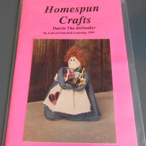 May include: A pink brochure with the title "Homespun Crafts" and a photo of a handmade doll. The doll has red hair, a white apron, and a blue plaid cape. The brochure is in a clear plastic sleeve.