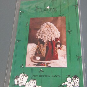 May include: A craft pattern packet featuring a Santa Claus doll. The packet has a green background with white illustrations of gifts and a snowman. The Santa doll has a red coat, white beard, and a burlap sack. Text on the packet reads "PLAIN JANE CREATIONS" and "#119 BUTTON SANTA."