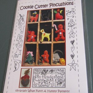 May include: A package featuring a pattern for making Cookie Cutter Pincushions. The design includes various felt pincushions shaped like gingerbread men, dogs, and other festive shapes. The text reads "oh-la-lah! What Fun!!! A Yummy Pattern!"