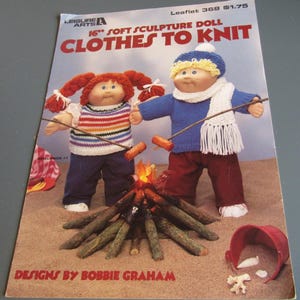 May include: A vintage soft sculpture doll pattern book titled "Clothes to Knit." The cover features two dolls, one with red pigtails and the other with a blue hat, standing near a campfire. The book is designed by Bobbie Graham.