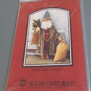 May include: A red card featuring a Santa Claus doll with a long white beard, holding a sack and a small Christmas tree. The text "SINT NIKKLAAS" is above the Sugar Camp Road logo.