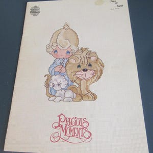 May include: A cross-stitch pattern booklet titled Precious Moments featuring a design of a child with blonde hair in a blue outfit, a white lamb, and a brown lion. The cover includes the text Peace on Earth, Book PM-4, and Designs by Gloria & Pat.