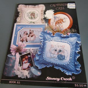 May include: A book cover titled "Captivating Cats" featuring cross-stitch designs. The cover displays various cat-themed needlework, including pillows and framed pictures. The designs showcase cats in different poses and settings, with a blue and white color scheme.