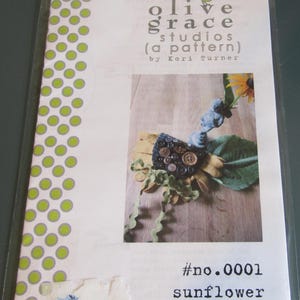 May include: A craft pattern package for a sunflower needle case. The package features a photo of a handmade sunflower with button details and green leaves. The text on the package reads "olive grace studios (a pattern) by Kori Turner" and "#no.0001 sunflower needle case."