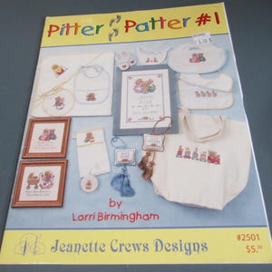 May include: A collection of baby-themed cross-stitch patterns, including bibs, framed pictures, and a tote bag. The designs feature teddy bears and the text "Pitter Patter #1" and "Jeanette Crews Designs". The color palette includes white, blue, and brown.