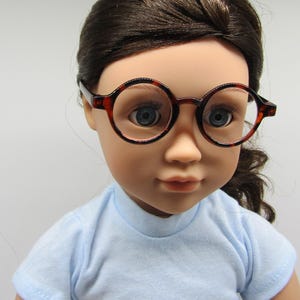 May include: A doll wearing a light blue shirt and round, brown-framed glasses.