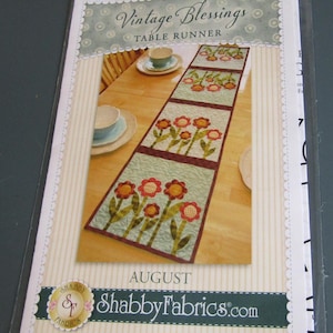 May include: A package featuring a floral table runner design. The table runner has a light blue background with red and orange flowers. The package is labeled "Vintage Blessings Table Runner" and "August". The brand name "Shabby Fabrics" is also visible.