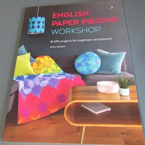 May include: A softcover book titled English Paper Piecing Workshop by Jenny Jackson, featuring a cover image of a living room with a colorful patchwork quilt, a round blue patterned pillow, and a wooden coffee table with a small potted plant.