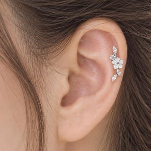 May include: A silver-toned ear cartilage piercing featuring a delicate floral design with white petals and small clear stones arranged in a vine-like pattern. The jewelry is positioned along the upper edge of the ear, showcasing a curved metal stem.