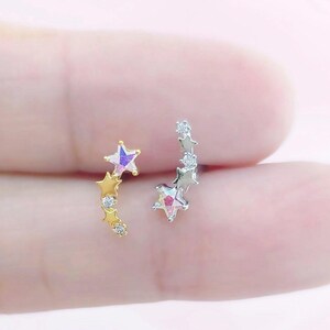 May include: Two star-shaped earrings. One earring is gold with three stars and small clear stones. The other earring is silver with three stars and small clear stones. The stars have a shimmering, iridescent finish.