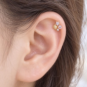 May include: A gold-toned cartilage earring featuring a cluster design with a small textured seashell, a five-pointed star with a clear stone, a round pink gemstone, and a tiny clear accent stone, all arranged together on a thin metal post.