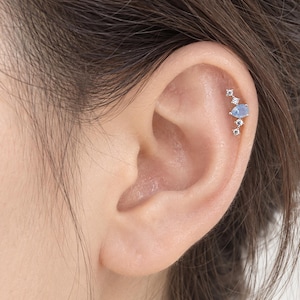 May include: A silver-toned cartilage earring featuring a central pear-shaped light blue stone flanked by four small clear round stones arranged in a vertical, curved line along the upper ear cartilage, secured with a thin metal post and backing.