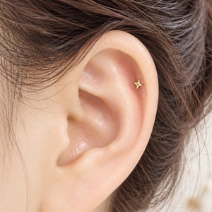 May include: A small gold-toned stud earring shaped like a four-pointed star. The jewelry piece is positioned in the upper cartilage area of the ear, featuring a polished metallic finish and a simple, symmetrical design with sharp, defined points.