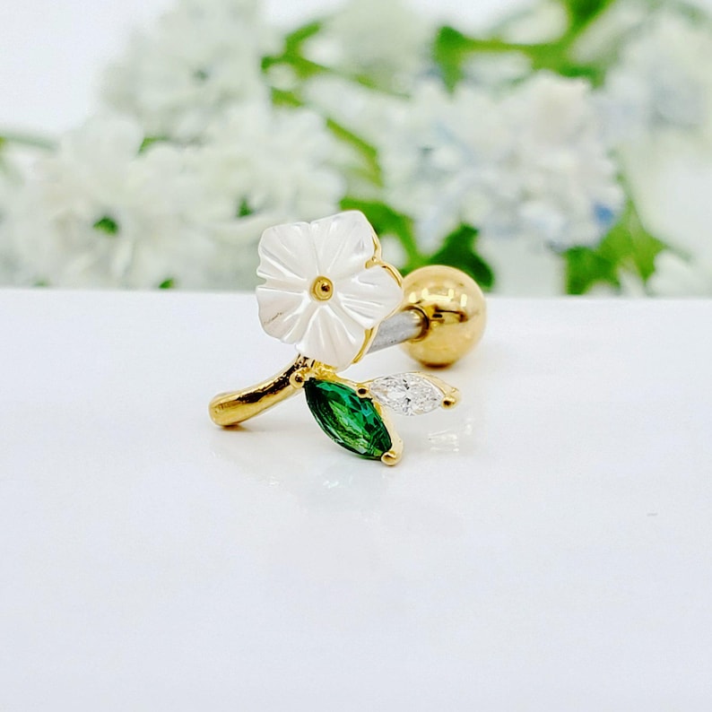 May include: A gold-tone barbell earring featuring a white flower-shaped charm, a marquise-cut green gemstone leaf, and a clear marquise-cut gemstone leaf. The jewelry piece includes a round gold-tone ball backing and a metallic post for attachment.