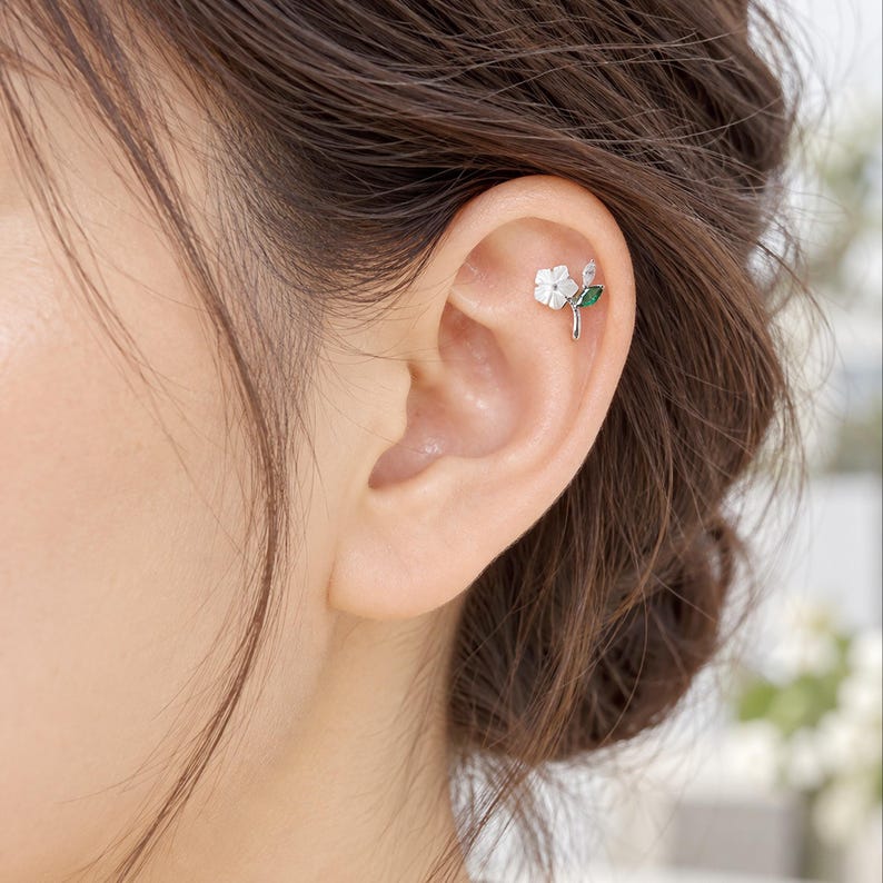 May include: A silver-toned ear stud featuring a small white flower with five petals, a single clear marquise-cut stone, and a single green marquise-cut stone, all arranged on a thin metal stem that curves along the upper cartilage of the ear.