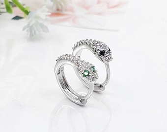 May include: A pair of silver hoop earrings with snake heads. One snake has a black stone for an eye and the other has a green stone.