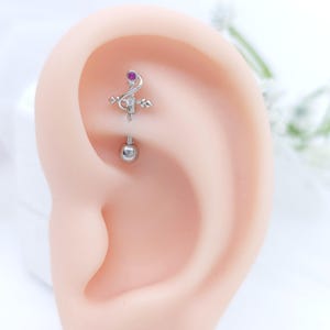 Op de afbeelding: Een witte keramische mok met een zwarte handgreep en de tekst "But First, Coffee" in een vetgedrukte, zwarte letter. De mok is gevuld met een donkere vloeistof, waarschijnlijk koffie, en staat op een lichtgekleurd oppervlak.