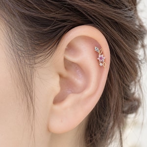 May include: A gold cartilage earring featuring a floral design with a central pink stone, surrounded by small clear stones and delicate gold leaf-shaped accents. The piece is positioned along the upper edge of the ear, showcasing a slender metal bar.