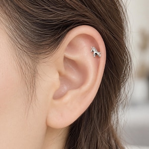 May include: A small, silver-toned unicorn stud earring positioned in the upper cartilage of an ear. The earring features a polished metallic finish and a distinct unicorn shape with a pointed horn, standing on four legs against the skin.