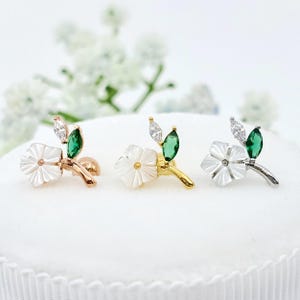 May include: Three stud earrings featuring a white carved flower, a marquise-cut clear stone, and a marquise-cut green stone. The earrings are set in rose gold, yellow gold, and silver-toned metal, each with a small post backing visible.