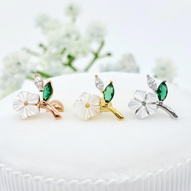 May include: Three stud earrings featuring a white carved flower, a marquise-cut clear stone, and a marquise-cut green stone. The earrings are set in rose gold, yellow gold, and silver-toned metal, each with a small post backing visible.