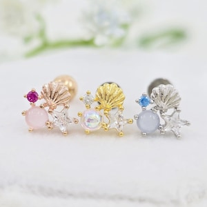 Op de afbeelding: Drie oorknopjes met een schelp, een ster en twee ronde stenen. De linker oorknop is roségoud met een roze steen, de middelste is goud met een iriserende steen en de rechter is zilver met een lichtblauwe steen en heldere kristallen.