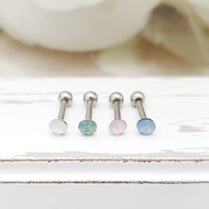 May include: Four silver-toned barbell earrings with colorful, faceted, round gemstones. The gemstones are in shades of white, teal, pink, and light blue. The earrings are displayed on a white surface.