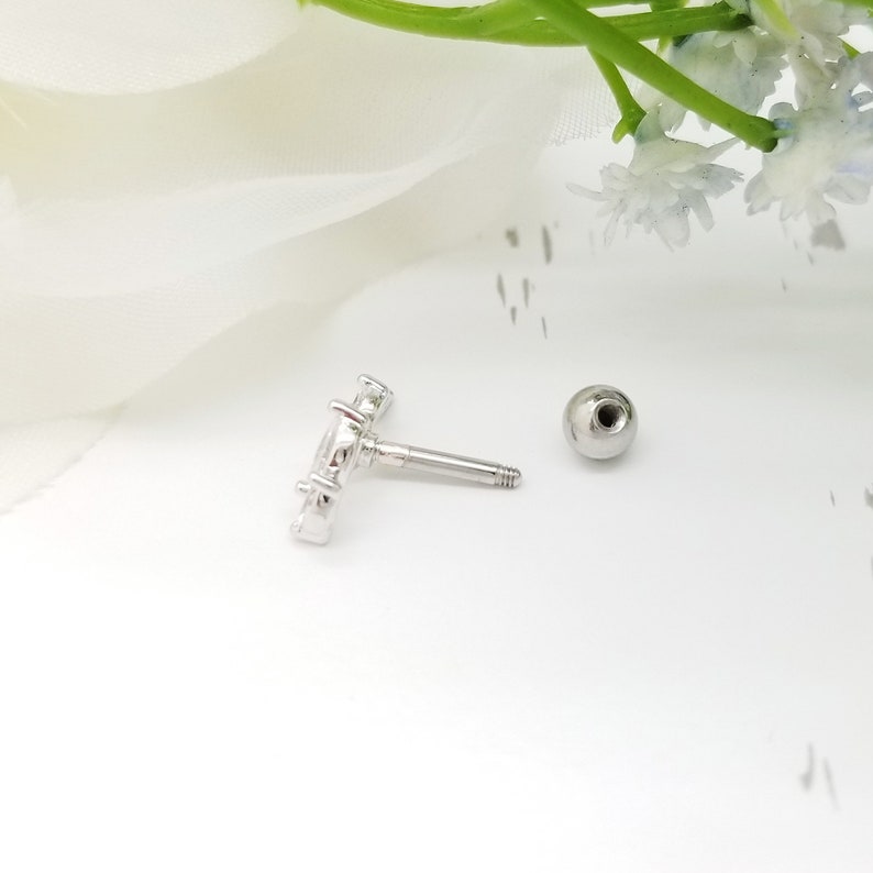 May include: A silver body jewellery piece with a clear crystal design. The jewellery is a barbell with a ball on one end and a crystal design on the other end.